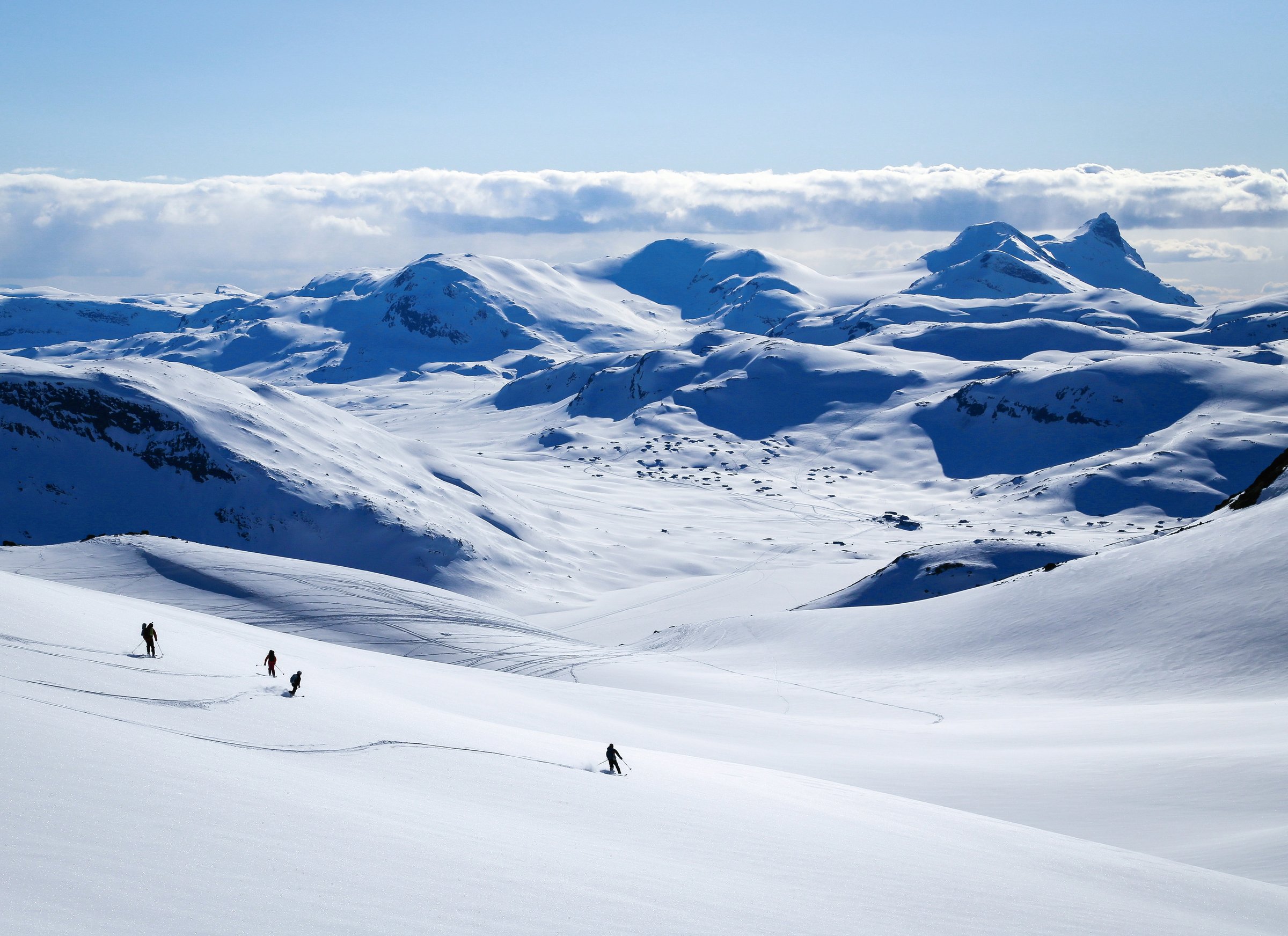 Fire skikjørere kjører ned en vid, snødekt fjellside fra Galdeberget, med utsikt mot Bygdin, snødekte fjell og et skydekke i horisonten.