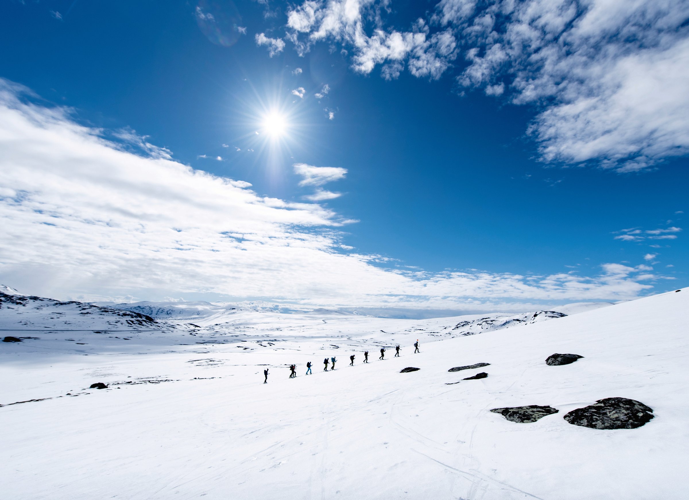 En lang rekke toppturløpere går på ski over snødekt fjell mot Høgdebrotet i Sørøst-Jotunheimen, under blå himmel med sol og drivende skyer.