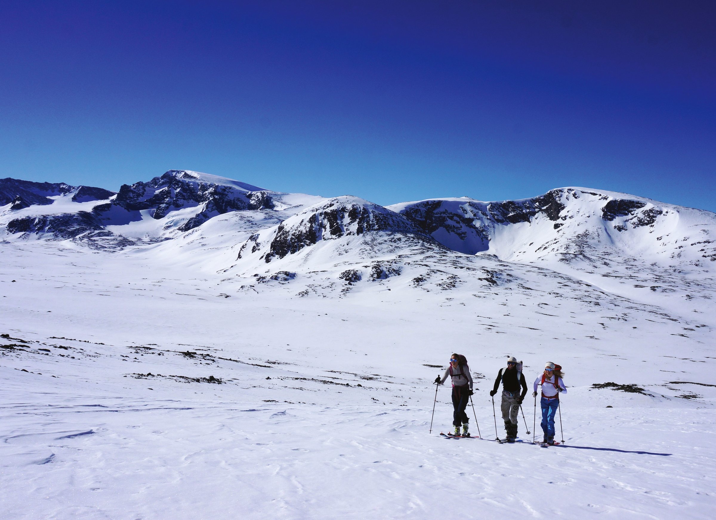 Tre skikjørere går på ski over et snødekt høyfjellsplatå fra Spiterstulen mot Glittertinden under klar blå himmel, med de snødekte toppene Skauthøe, Spiterhøe og Leirhøe i bakgrunnen.