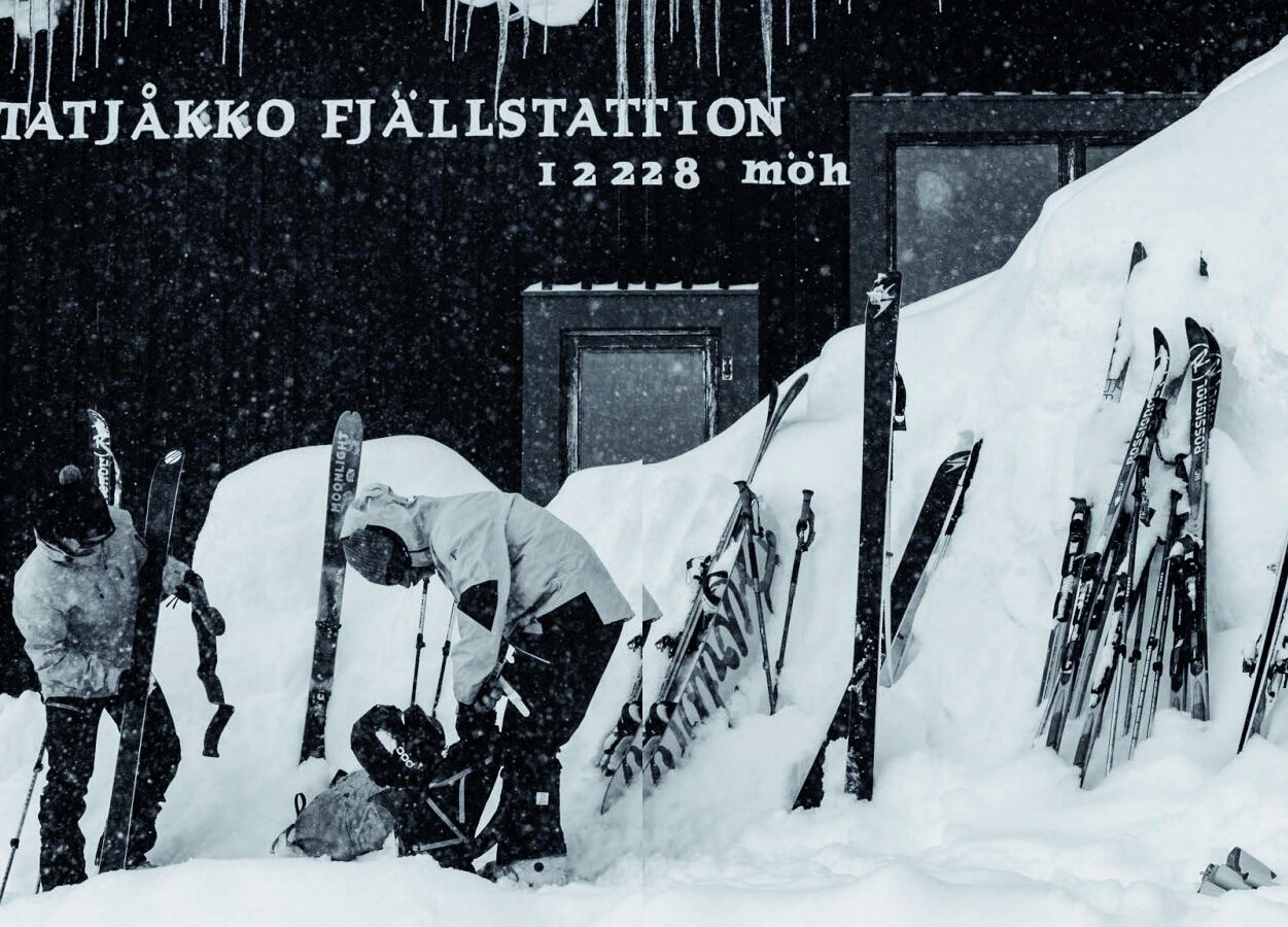 Svart-hvitt bilde av ski stukket ned i snøen utenfor Låktatjåkka Fjällstation, med skikjørere som gjør seg klare i snøværet.