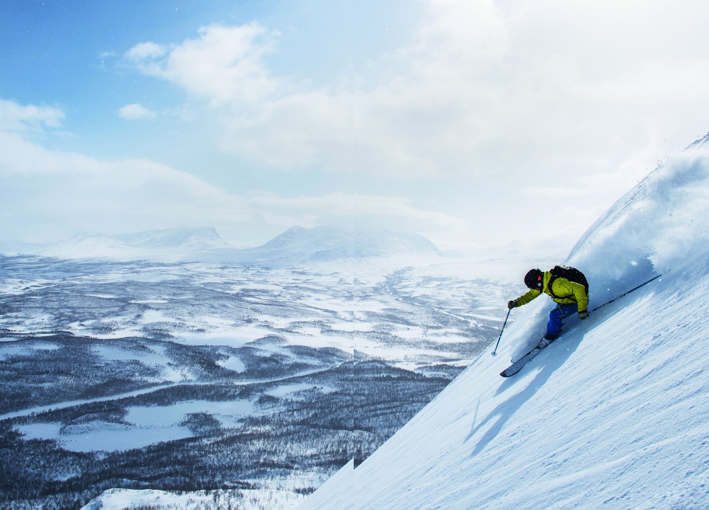 Skikjører i gul jakke svinger ned en bratt snøhelling på Slåttatjåkka, med Abiskodalen og Lapporten i bakgrunnen.