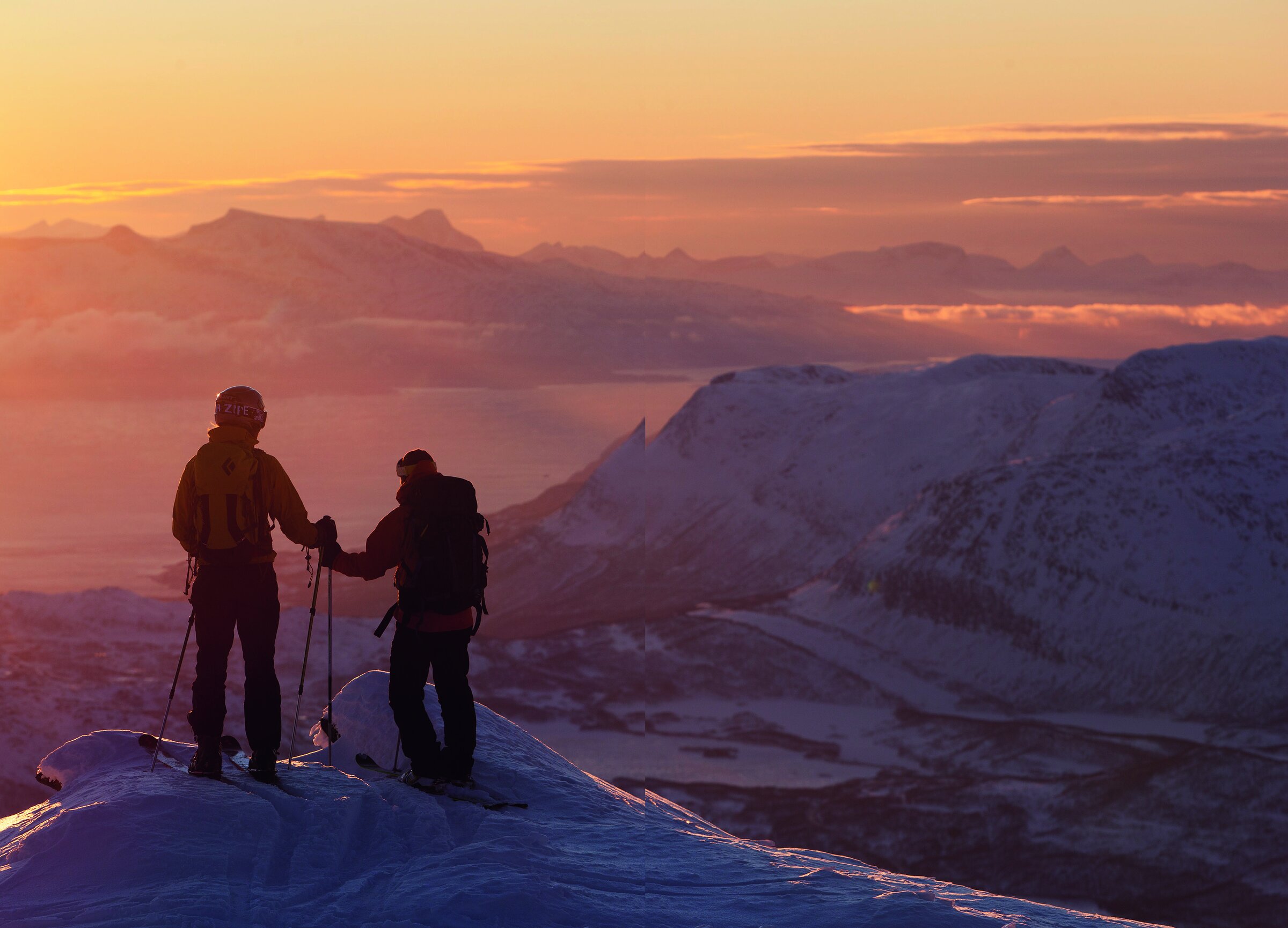 To skikjørere står i silhuett på toppen av Spanstinden i oransje kveldslys, med fjell og fjord i bakgrunnen.