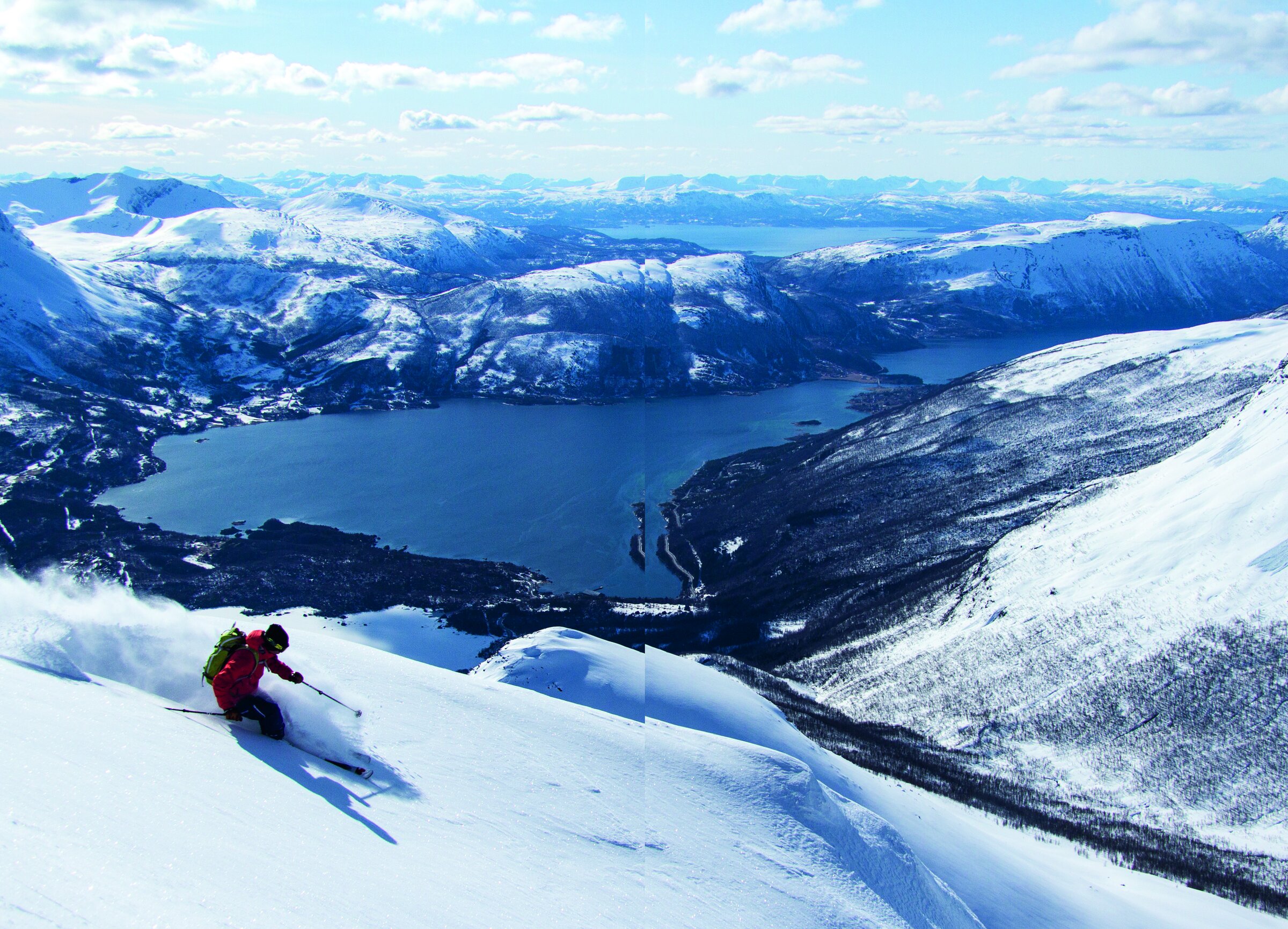 Skikjører i rød jakke svinger ned en snødekt fjellside på Oldertinden, med Grovfjorden og snødekte fjell i bakgrunnen.