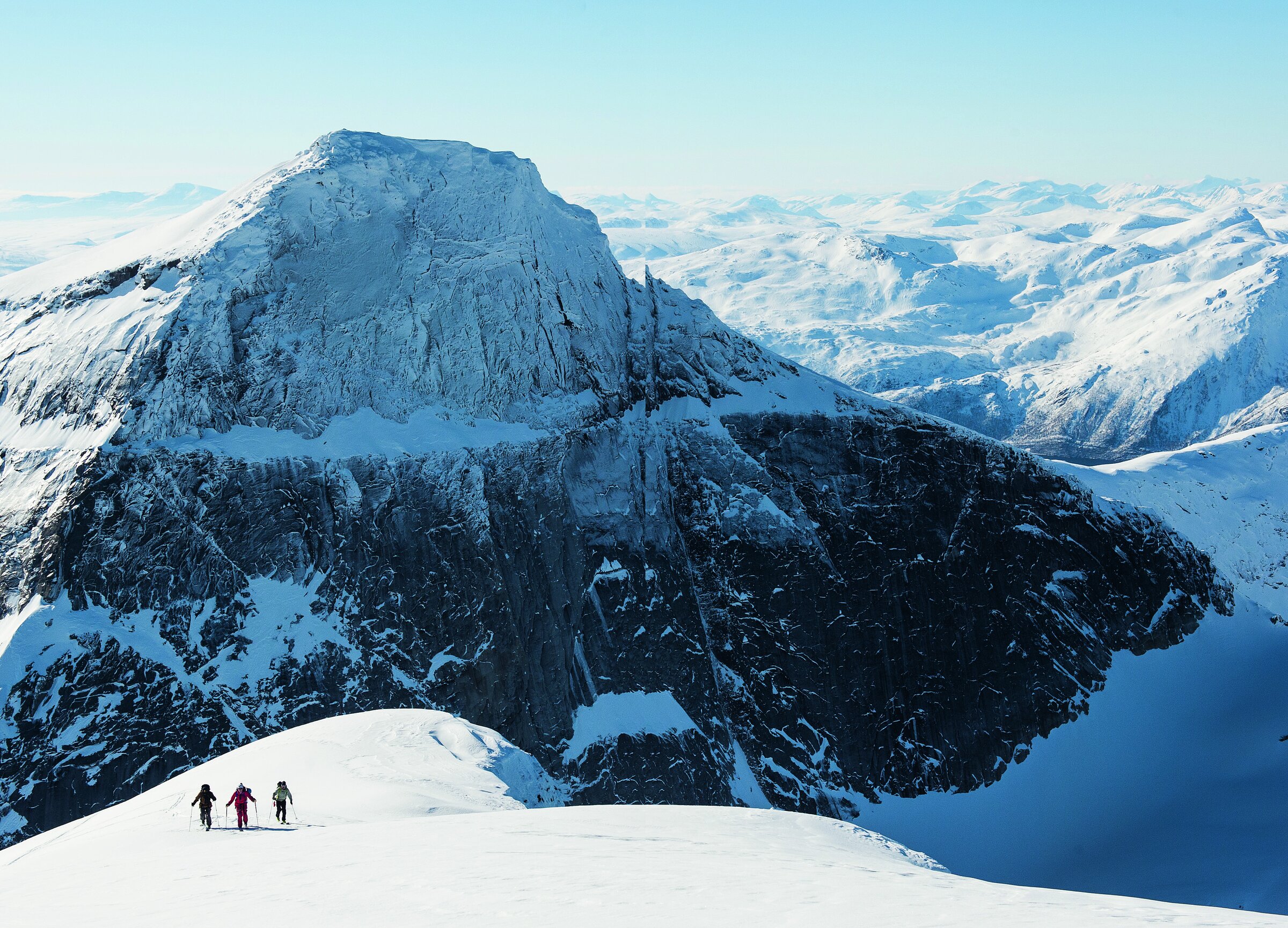 Tre skikjørere går opp en snødekt rygg mot Stetinds bratte granittvegger, med fjell og fjorder i bakgrunnen.