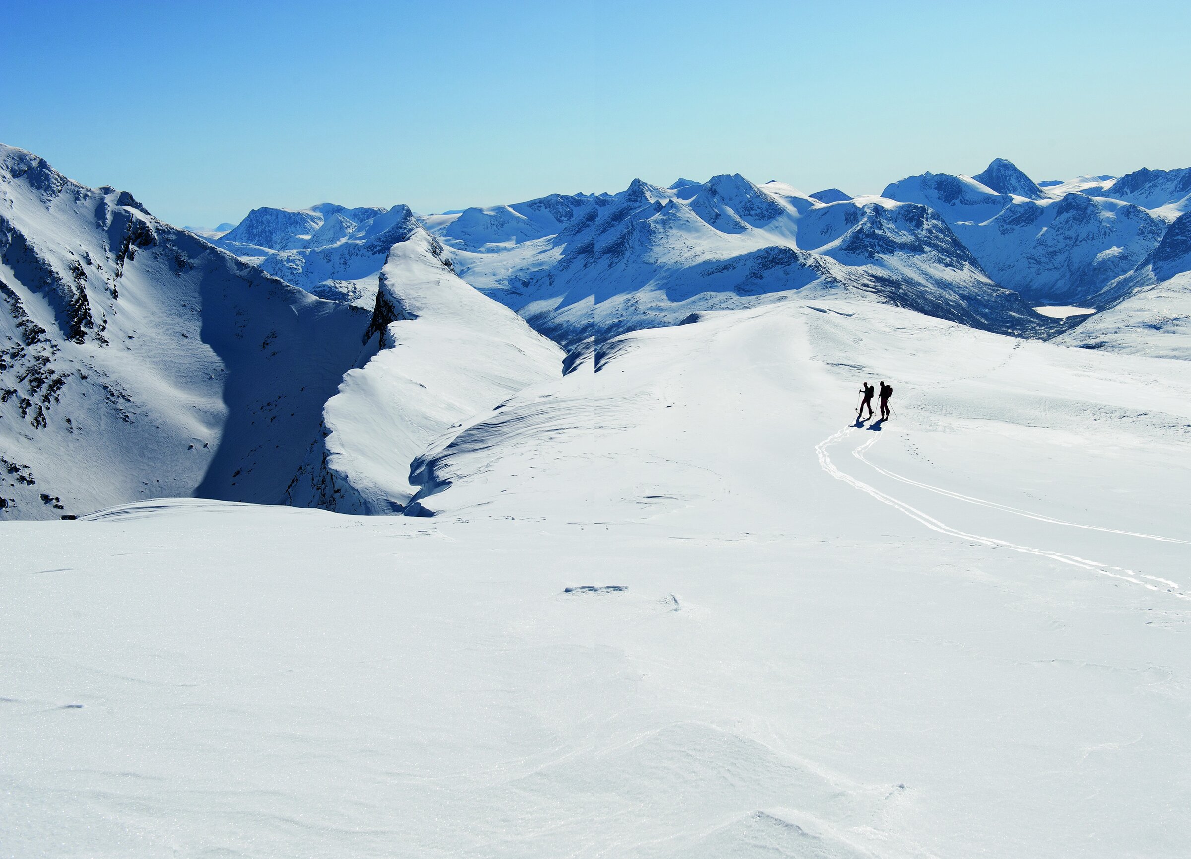 To skikjørere på et stort snødekt høyfjellsplatå, med spisse alpine topper rundt Narvik i bakgrunnen.