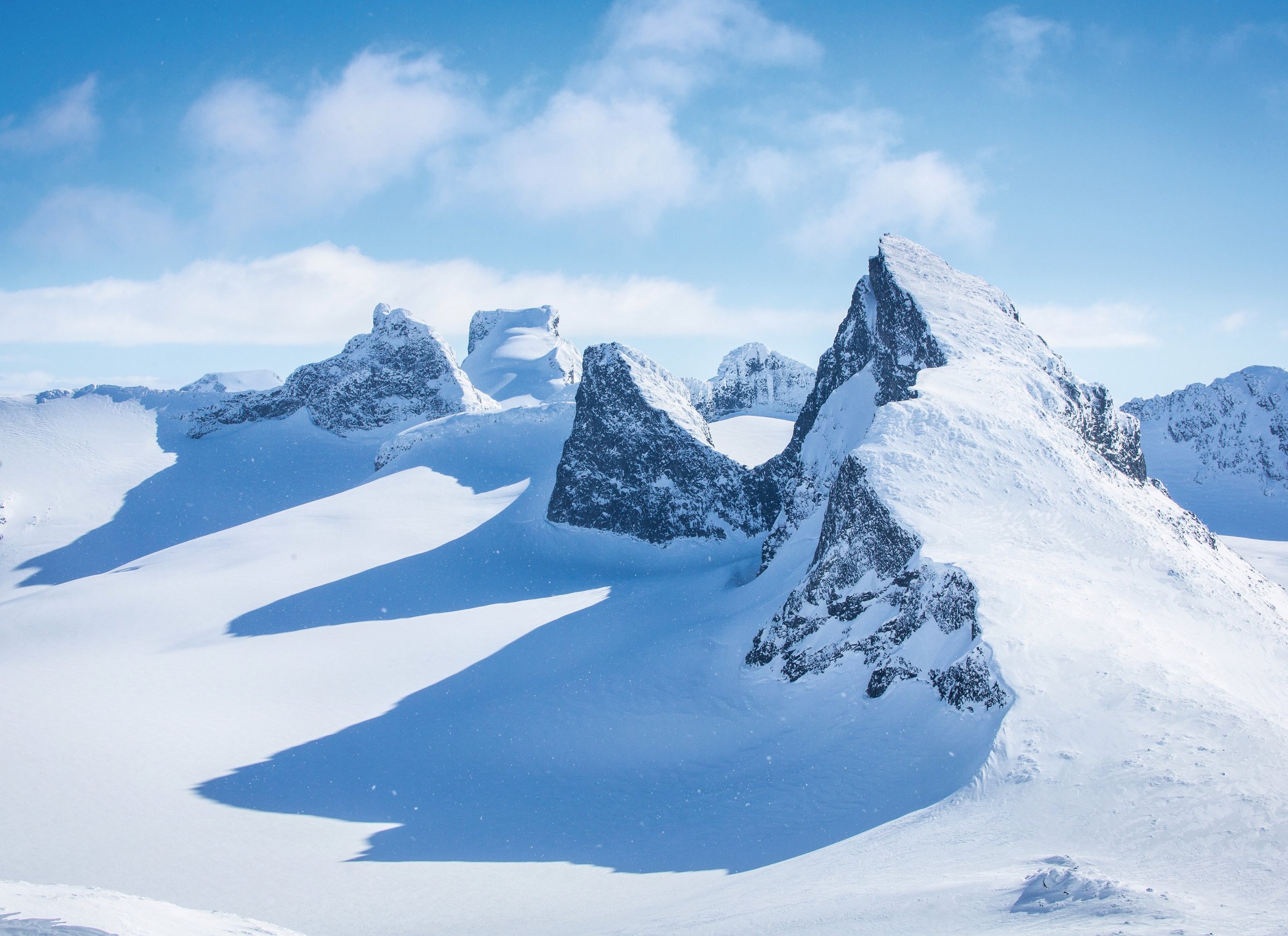 Spisse, snødekte tinder stikker opp av en bre med lange skygger. Utsikt fra Veslbreatinden mot Storbrean og Smørstabbtindane i Jotunheimen.